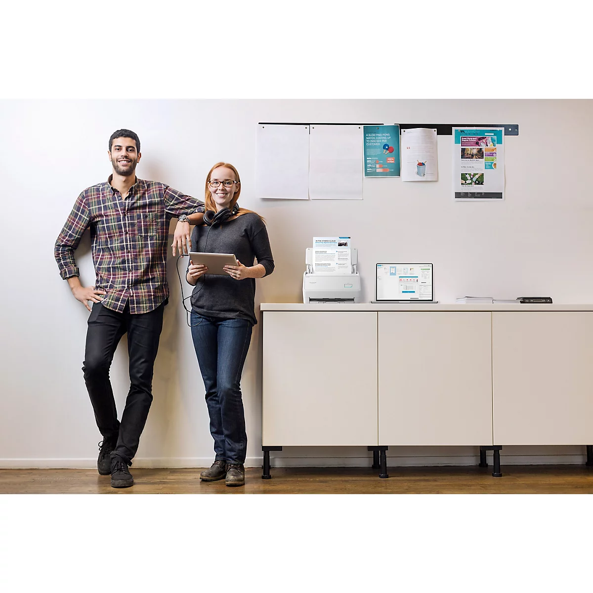 Ein Mann und eine Frau lächeln neben einem modernen Drucker und Laptop in einem hellen Büro.