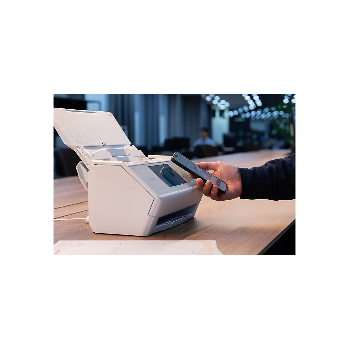 Eine Person hält ein Smartphone über einen weißen Dokumentenscanner auf einem Holztisch in einem Büro.