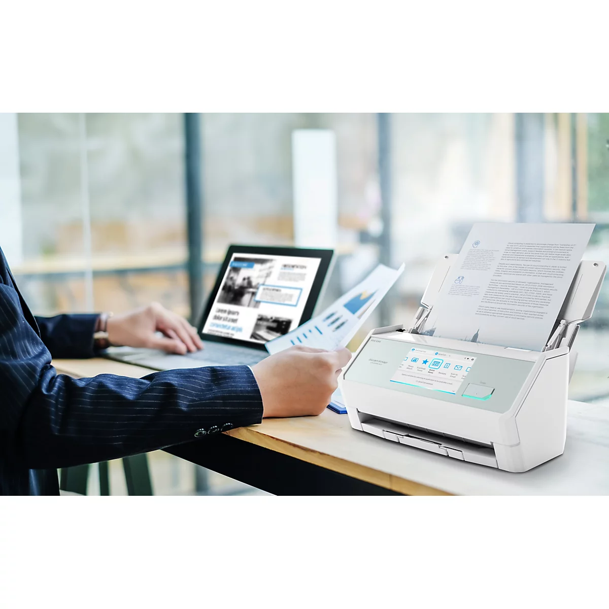 Ein moderner Dokumentenscanner steht auf einem Bürotisch neben einem Laptop, während eine Person mit Papieren arbeitet.