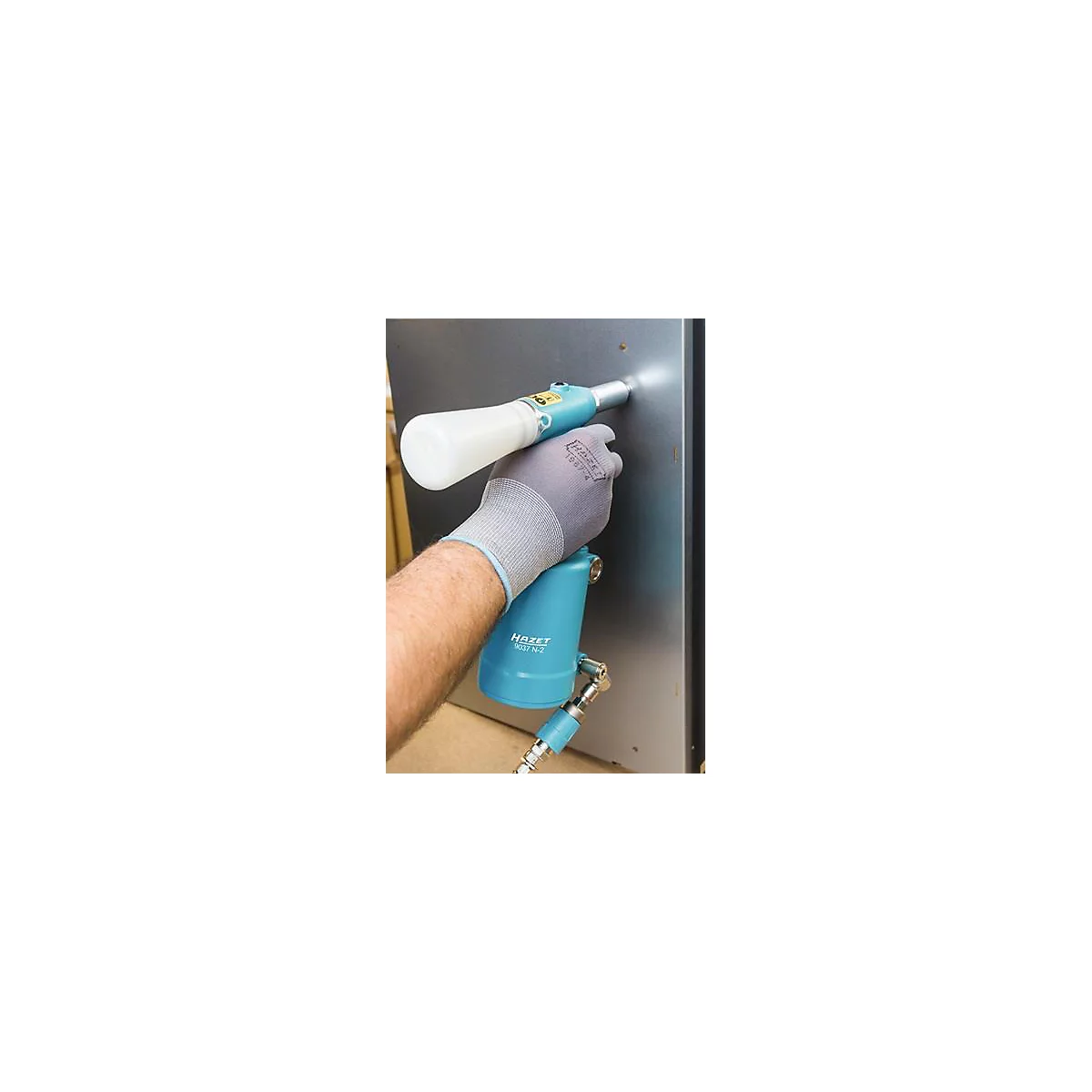 Une personne tient un outil pneumatique bleu contre une plaque métallique. Le gant porte le logo HAZET. Le fond est flou.