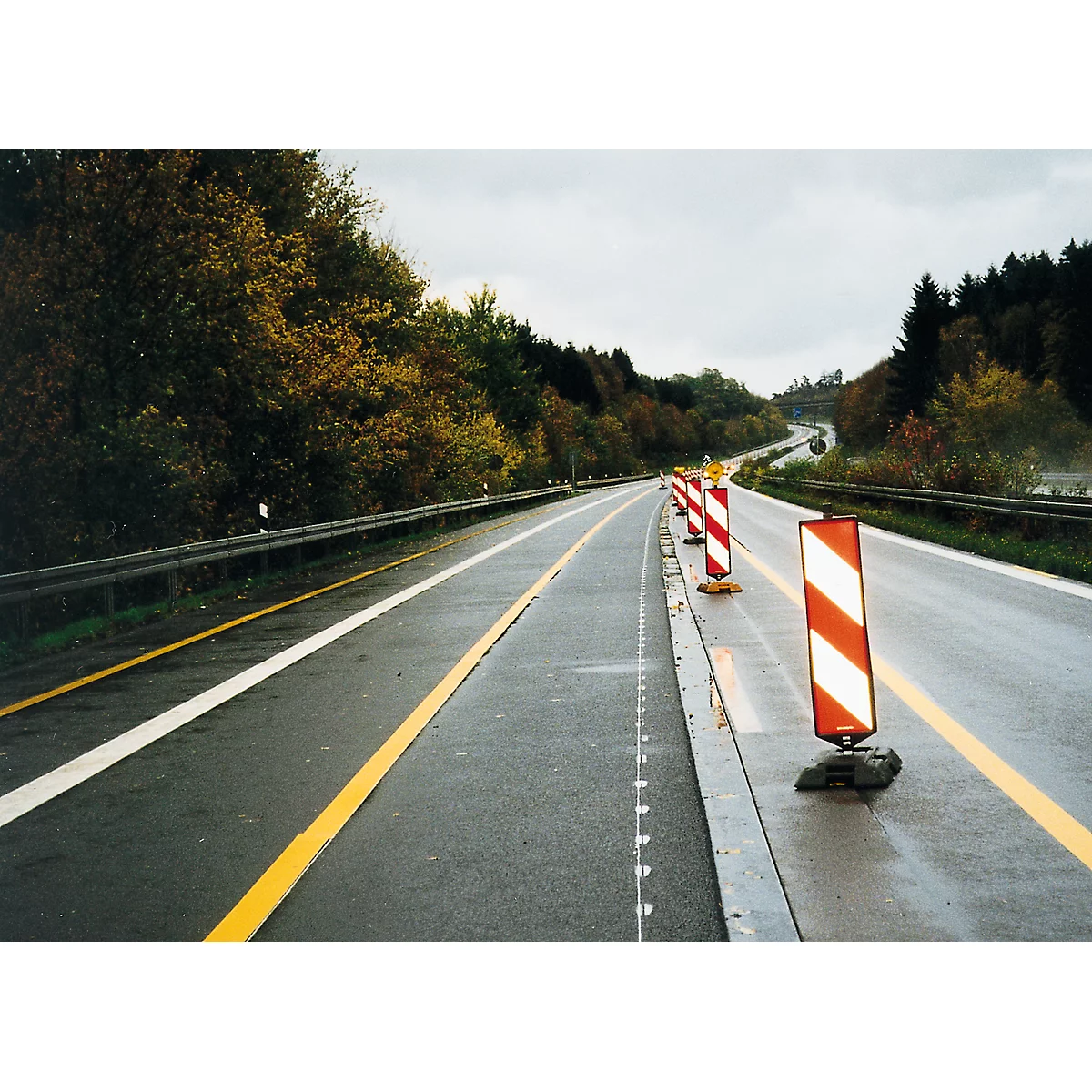 A two-lane highway with red and white warning beacons on a curve. There are trees on the roadside, the sky is overcast.