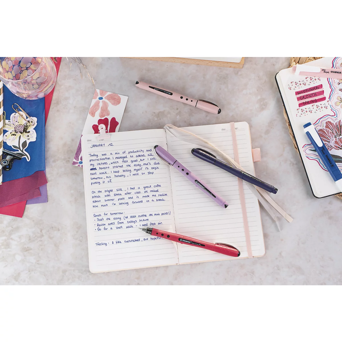 Gros plan sur un carnet, des stylos et des accessoires. Divers stylos reposent sur le carnet ouvert avec du texte.