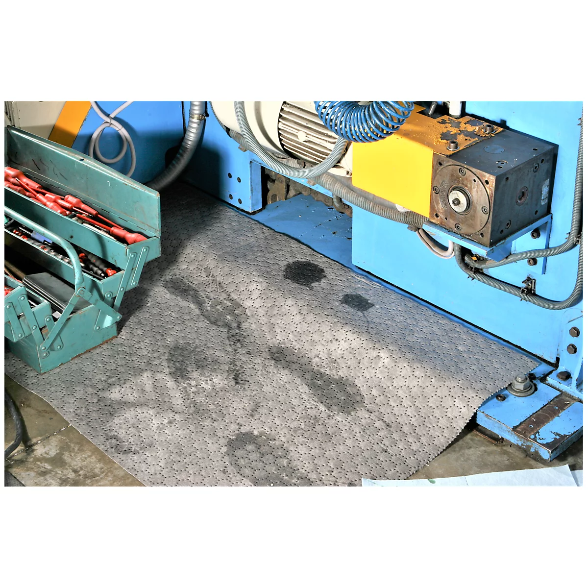 Alfombra de trabajo gris hexagonal con manchas de aceite, frente a una máquina azul y una caja de herramientas.