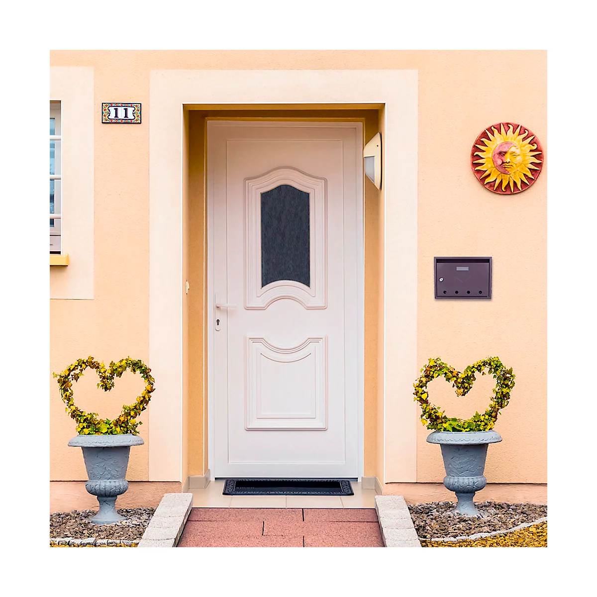 Porte d'entrée blanche avec insert en verre, au-dessus le numéro de la maison. À gauche et à droite, un pot avec des plantes en forme de cœur.