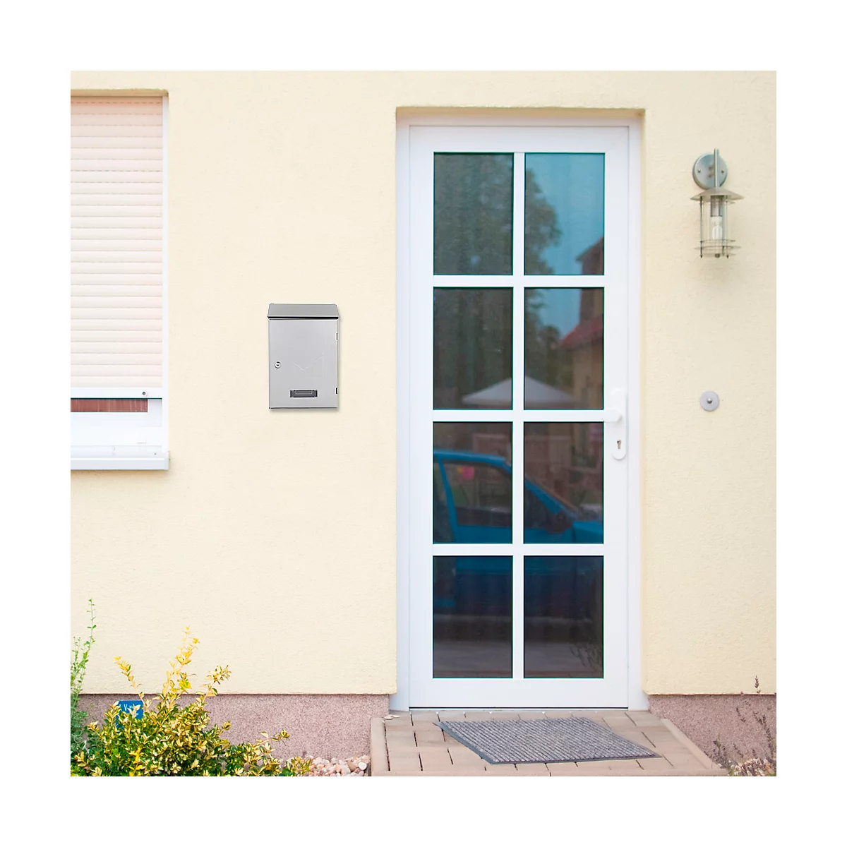 Boîte aux lettres grise sur un mur de maison jaune. Une porte et une fenêtre blanches, ainsi qu'une lampe extérieure sont également visibles.