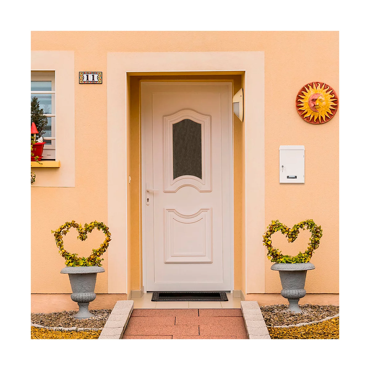 Porte d'entrée blanche avec une fenêtre, flanquée de jardinières en forme de cœur. Au-dessus de la porte, une boîte aux lettres et une peinture murale. Le mur de la maison est orange.