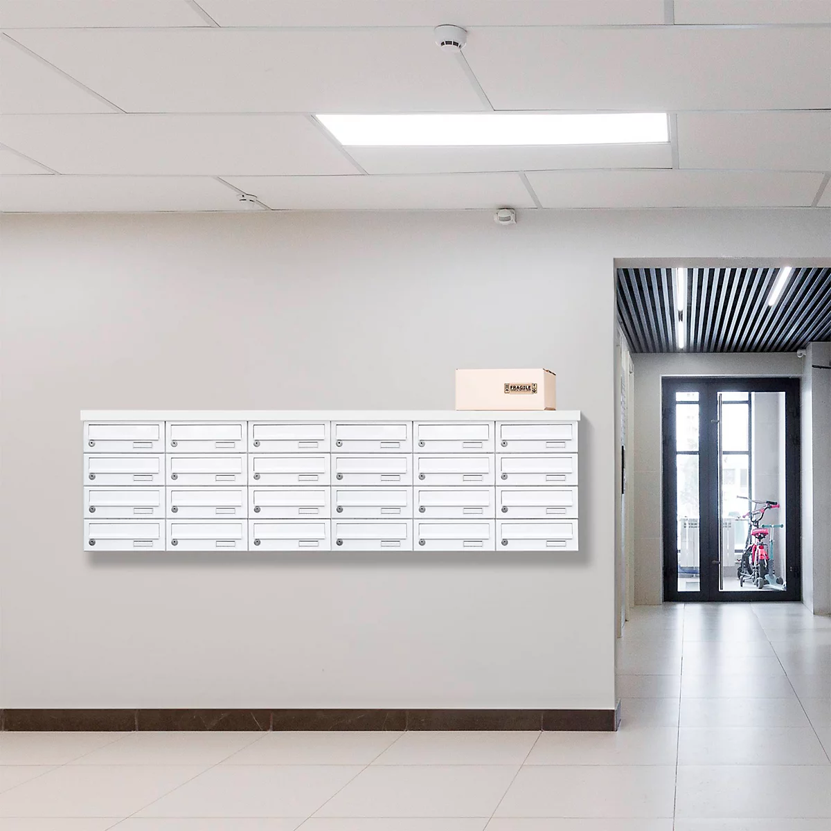 Reihenweise weisse Briefkästen an grauer Wand, über einem Flur mit Fenstern und einem Paket.