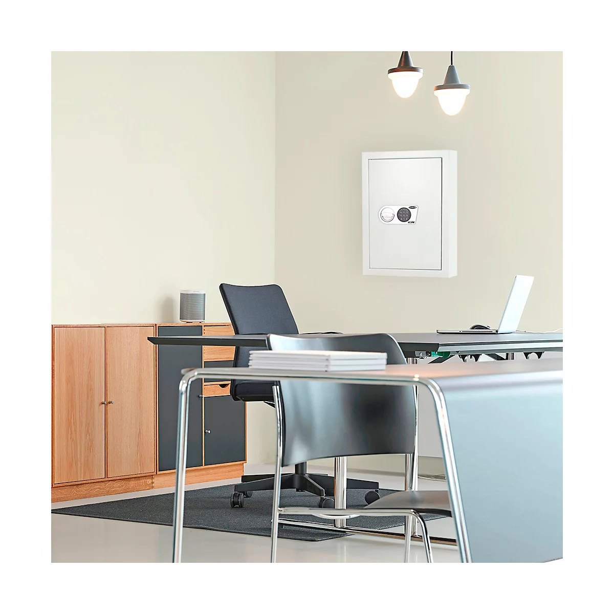 Un coffre-fort blanc est accroché au mur dans un bureau. Un bureau avec une chaise est placé devant le coffre-fort, une commode derrière.