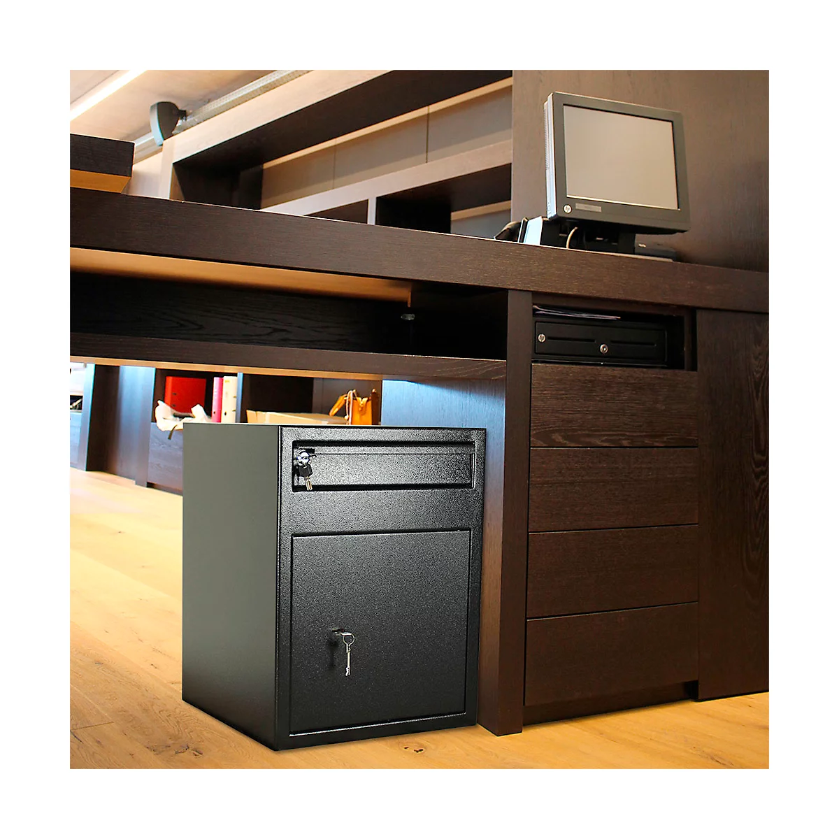 Un coffre-fort noir est placé sous un bureau. Un moniteur et des tiroirs sont également visibles.