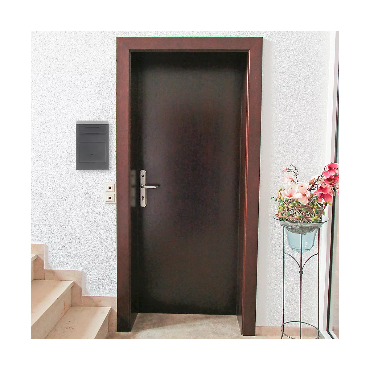 Une porte en bois foncé avec une poignée argentée. À droite, un présentoir à fleurs, et à gauche, un escalier.