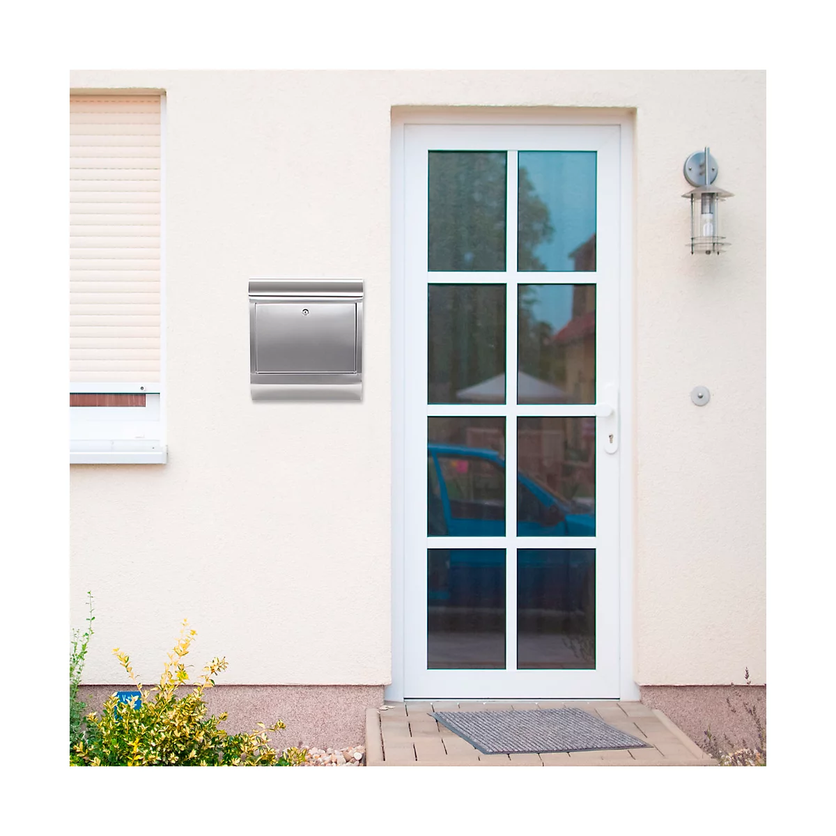 A silver mailbox on a light colored house wall next to a white door. A car can be seen in the background.