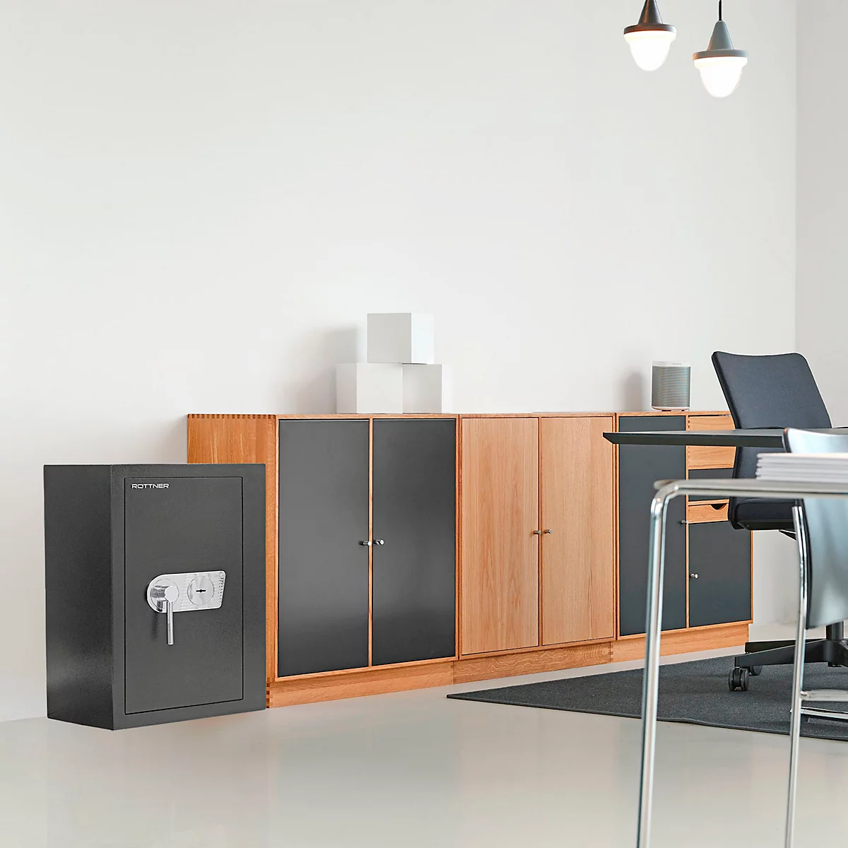 A gray safe stands next to a light wood cabinet in front of a white wall. Two lamps hang from the ceiling.