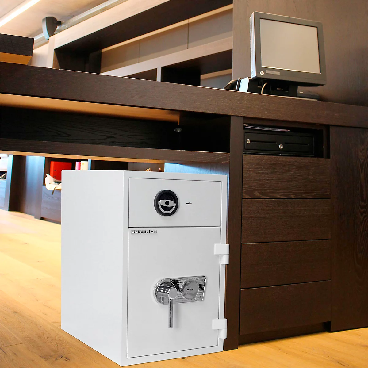 Weisser Tresor steht unter einem braunen Schreibtisch, daneben ein Computer-Monitor.