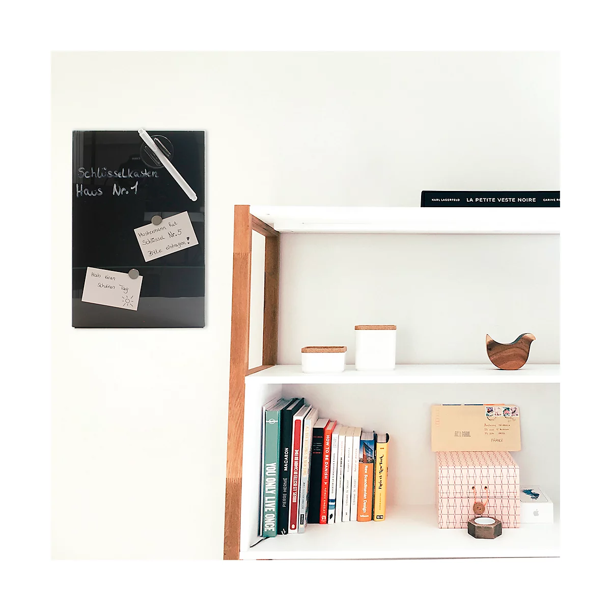 Zwart whiteboard met notities en pen op witte muur. Daarnaast een plank met boeken, decoratie en dozen.
