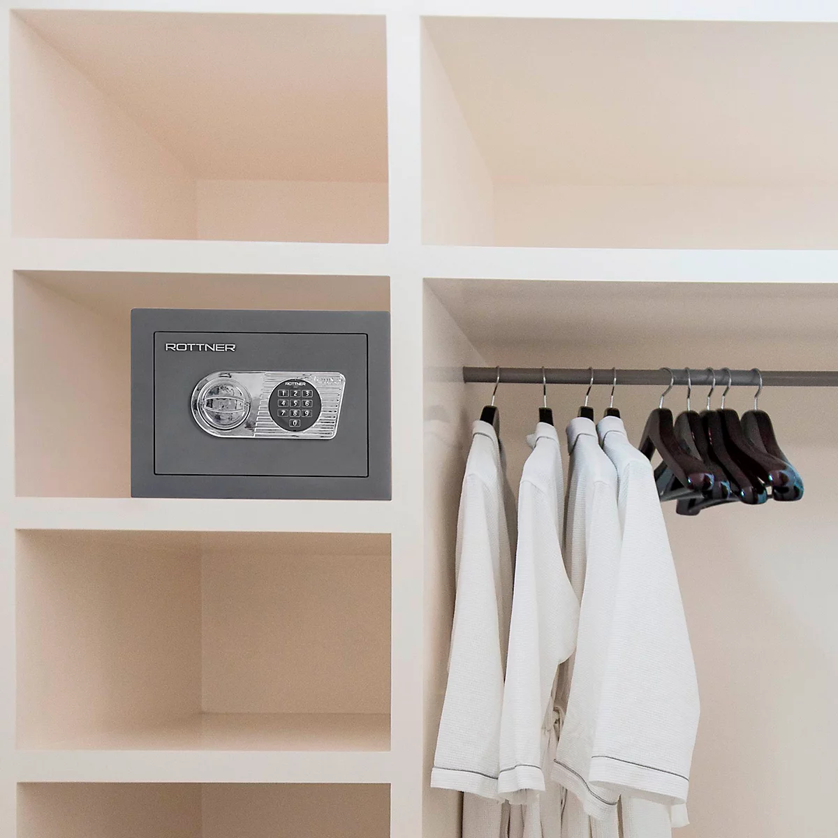 Grauer Tresor mit Zahlencode und Kleiderbügel mit weissen Bademänteln in einem Schrank. Rechts leere Kleiderbügel.