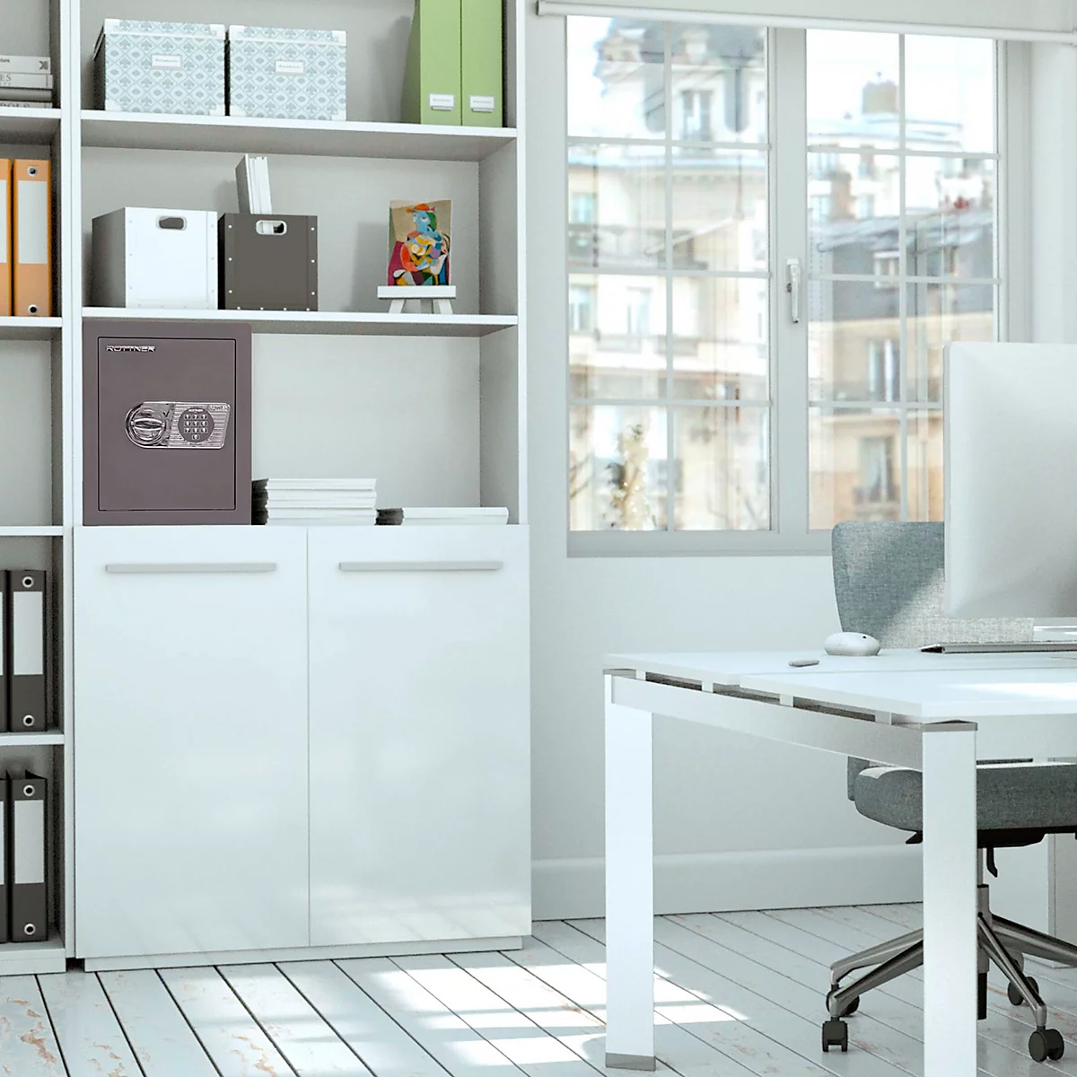 Ein Büro mit hellem Mobiliar, einem Tresor und einem Fenster mit Stadtblick. Am Schreibtisch steht ein Monitor.