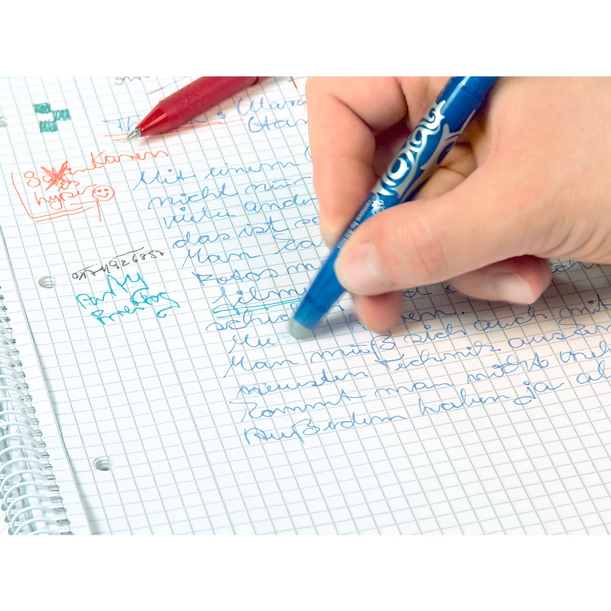 Mano borrando texto en papel cuadriculado con bolígrafo borrador azul. Un bolígrafo rojo está al lado.