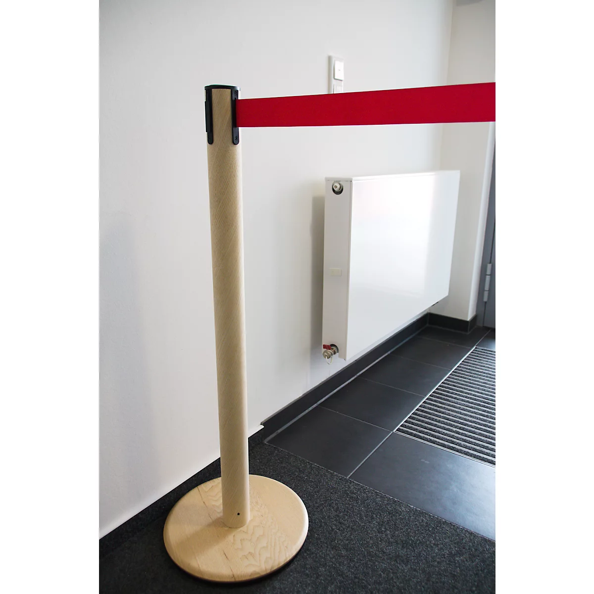 Un ruban rouge de barrière est attaché à un poteau en bois. En arrière-plan : radiateur, tapis gris et carreaux.