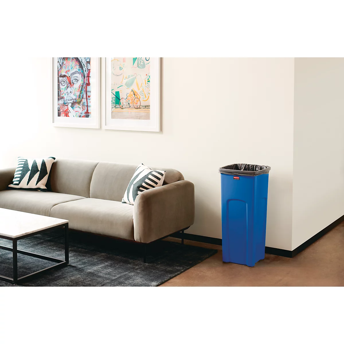 Blauer Mülleimer in einem Wohnzimmer. Neben einem Sofa, einem Tisch und Gemälden an der Wand.