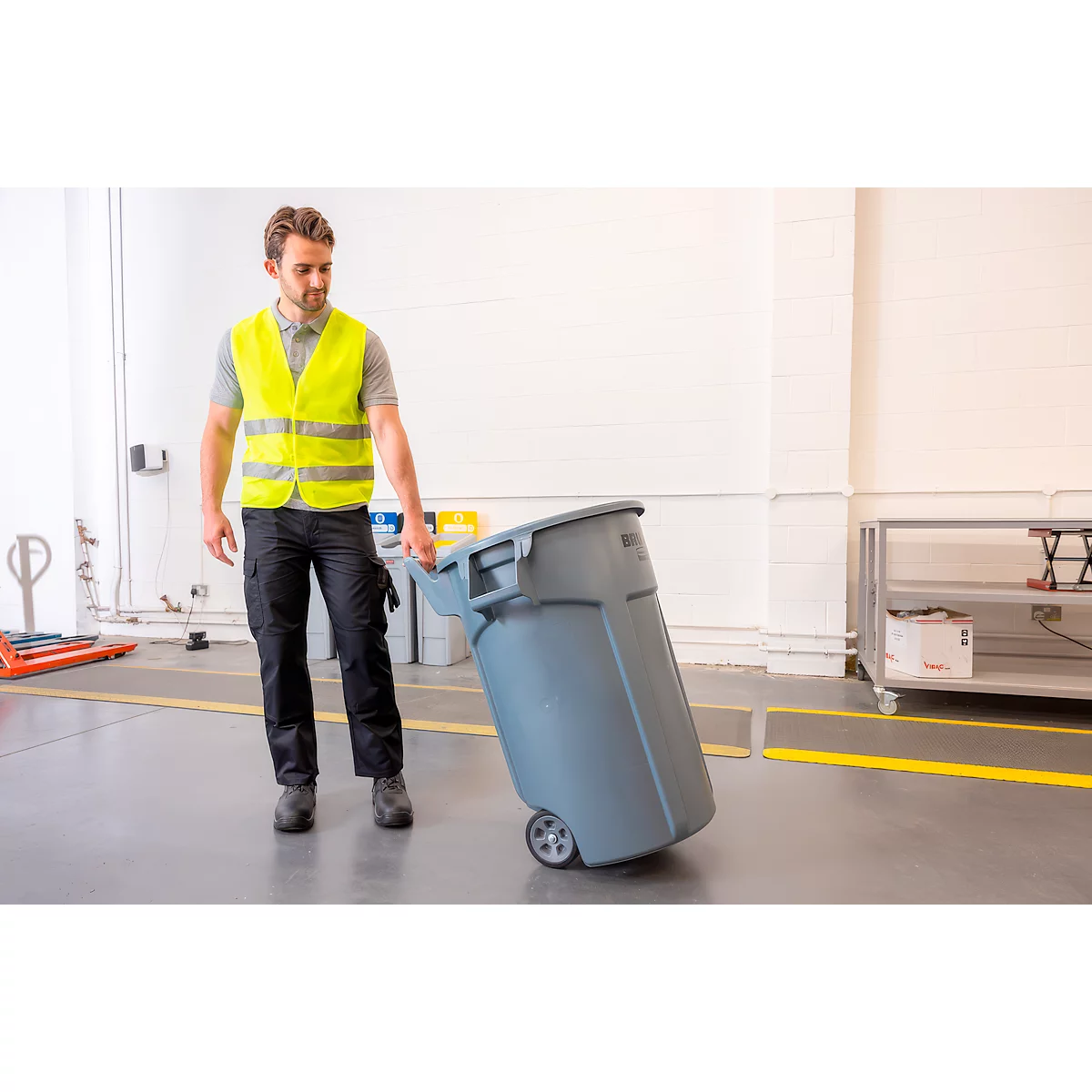 Ein Arbeiter in Warnweste zieht eine graue Abfalltonne auf Rollen durch ein Lager.