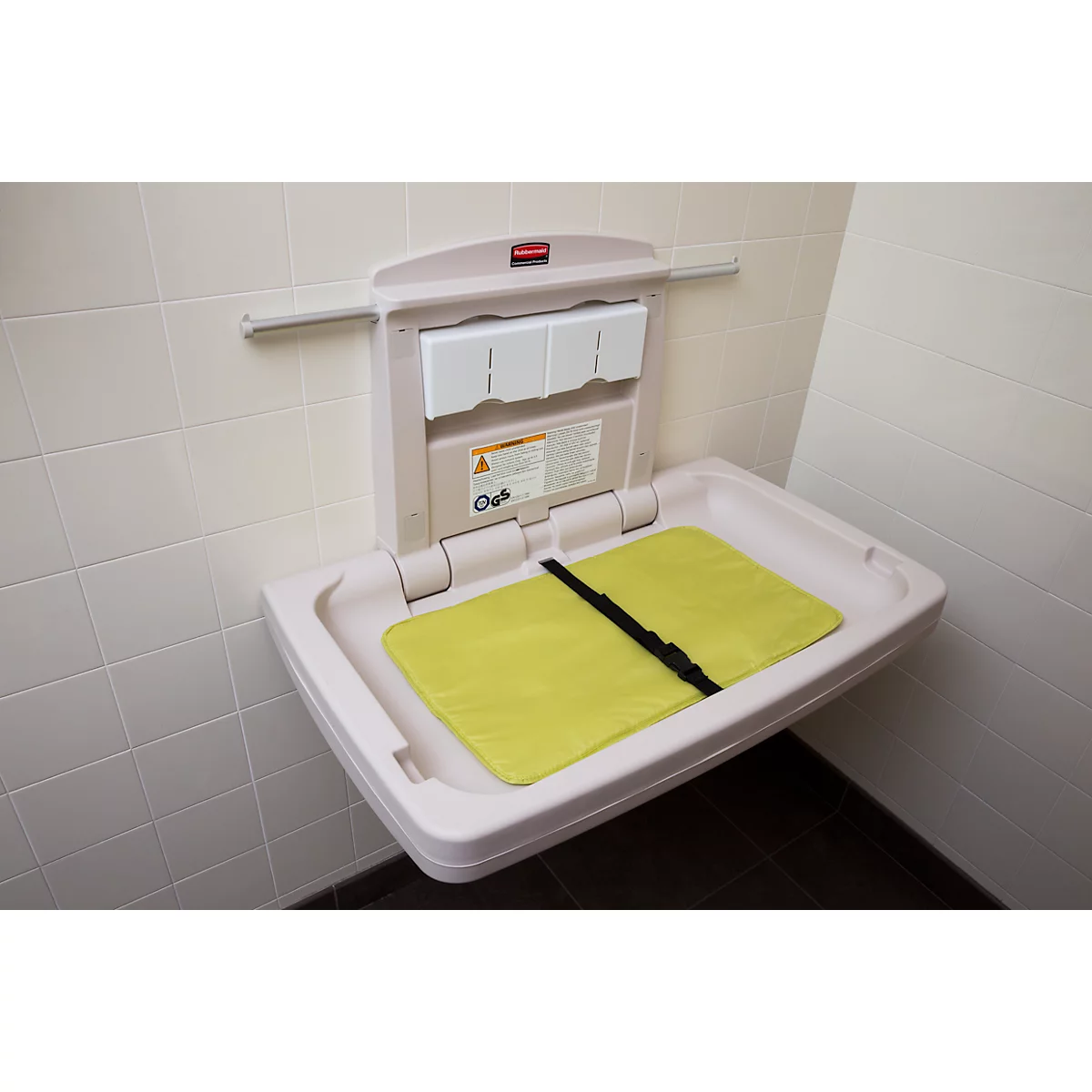 Aan de muur gemonteerde luiertafel, geopend. Gele mat, zwarte band, beige kast tegen witte muur. "Rubbermaid" rechtsboven.