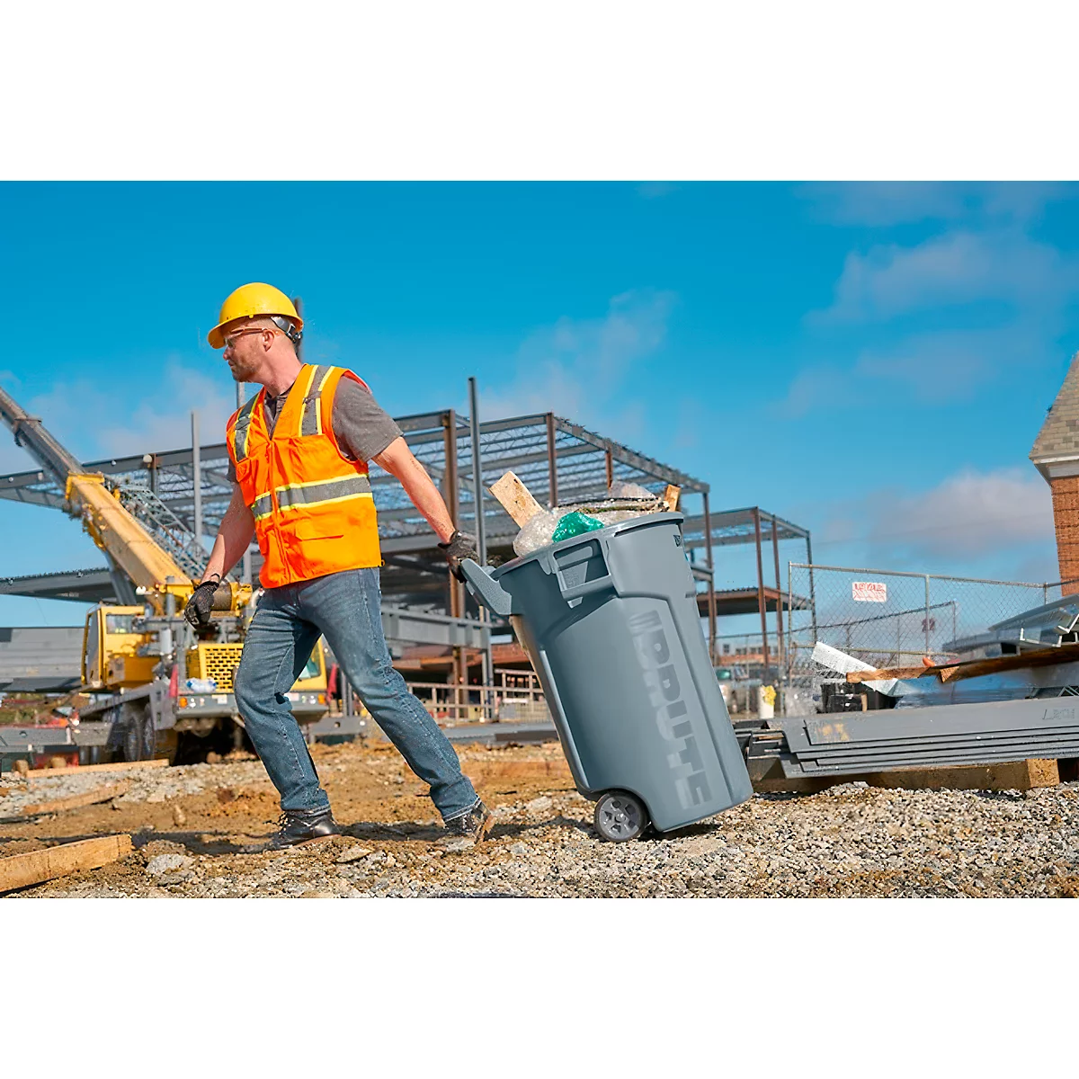 Un ouvrier du bâtiment avec un casque de sécurité tire une poubelle grise sur un chantier.