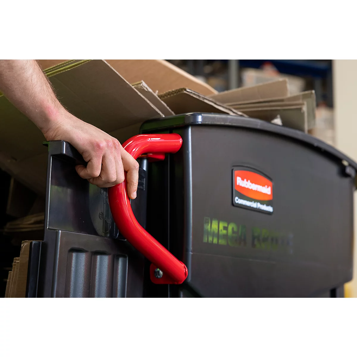 Close-up van een persoon die de handgreep vasthoudt van een zwarte afvalcontainer met een rode handgreep, gevuld met kartonnen dozen.