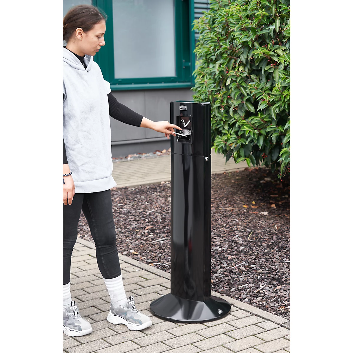 Junge Frau wirft etwas in eine schwarze Mülltonne. Sie trägt eine graue Kapuzenjacke und schwarze Hose.
