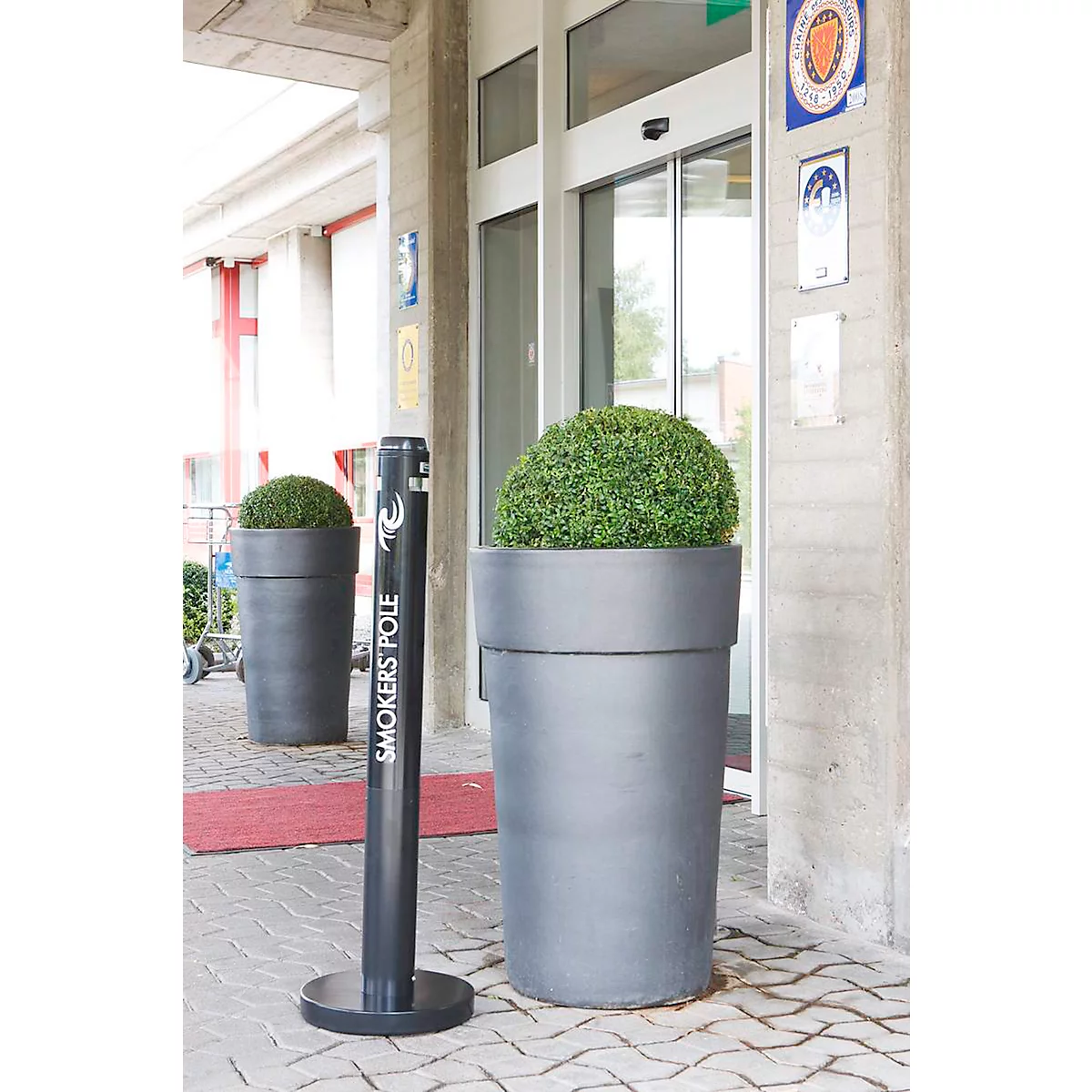 Dunkelgraue Kübel mit grünen Büschen flankieren eine Glastür. Ein schwarzer "Smokers Pole" steht davor.