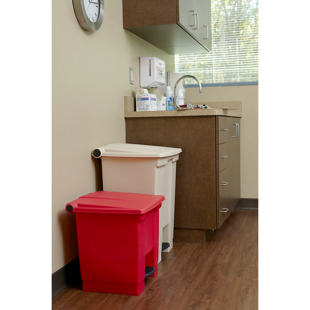 Roter und weißer Mülleimer im Behandlungsraum, Waschbecken und Uhr an der Wand.