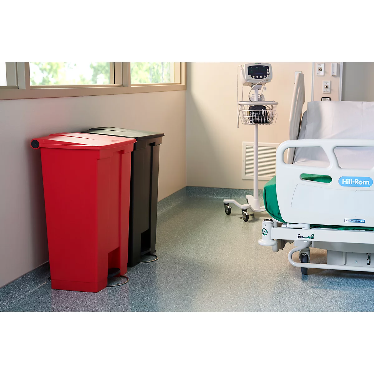 Roter und schwarzer Abfalleimer in einem Krankenhauszimmer, neben einem Bett und medizinischen Geräten.