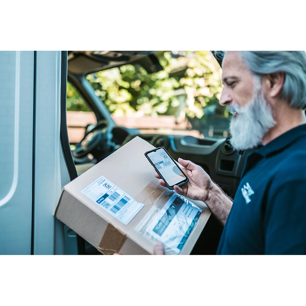 Ein Mann mit grauem Bart scannt einen Paketaufkleber mit einem Smartphone. Das Paket liegt im Laderaum eines Transporters.