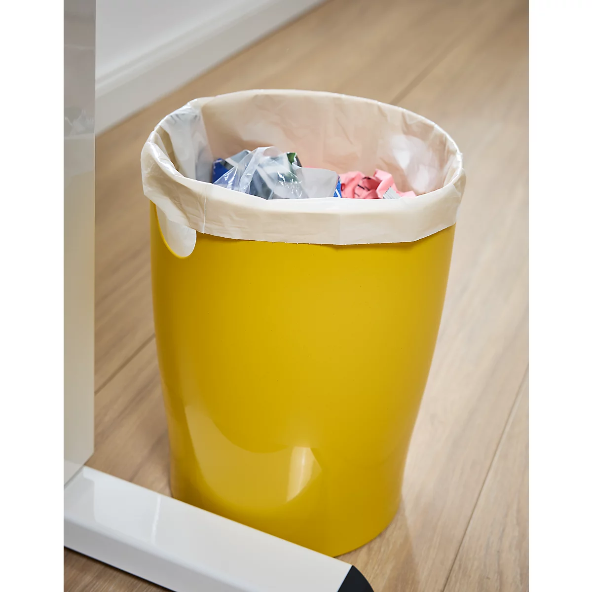 Poubelle jaune avec sac en papier rempli de déchets, posée sur un parquet.