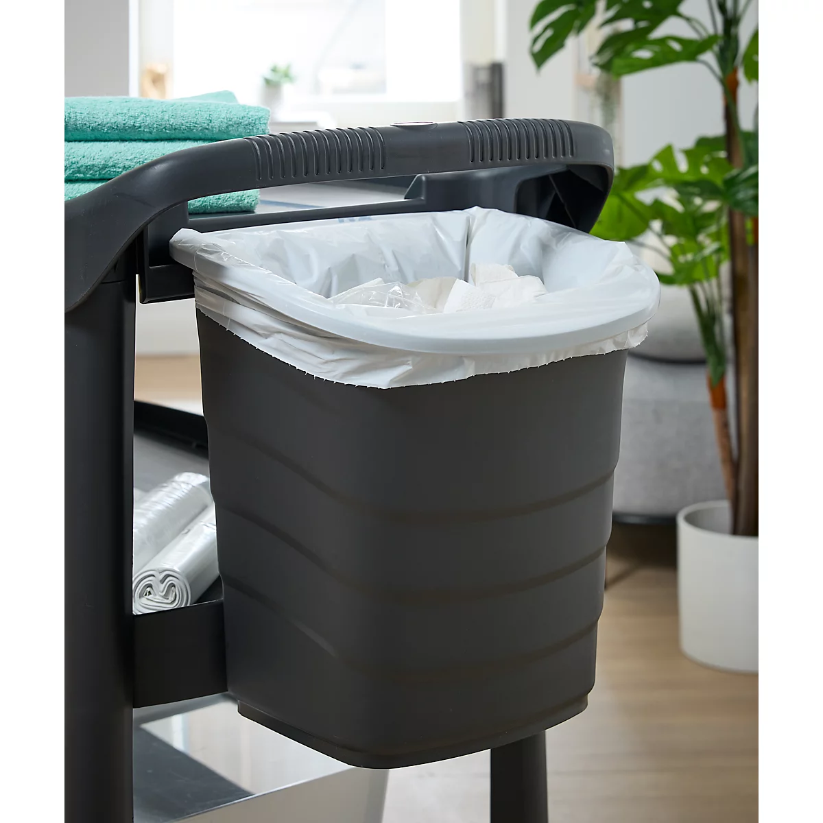 Poubelle grise avec sac poubelle blanc sur un chariot de nettoyage noir. En arrière-plan, une plante et une serviette verte.
