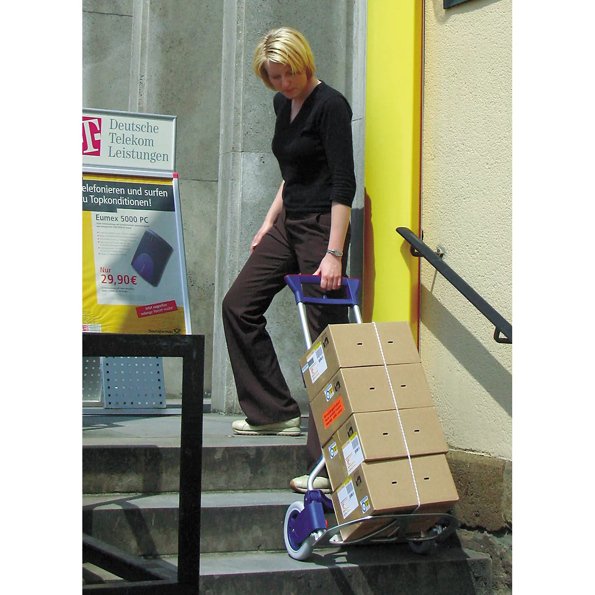Woman pulls a cart loaded with packages up stairs. In the background an advertisement sign with a telecom logo.