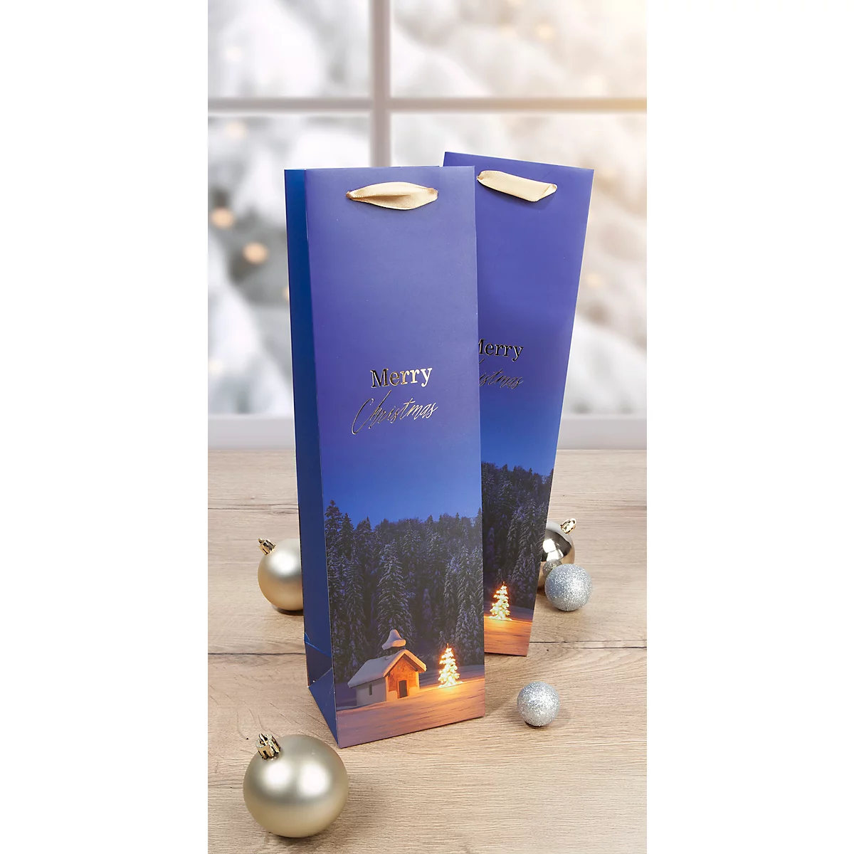 Deux sacs cadeaux de Noël bleus avec imprimé paysage et poignées dorées sont posés sur une table en bois. La neige scintille en arrière-plan.