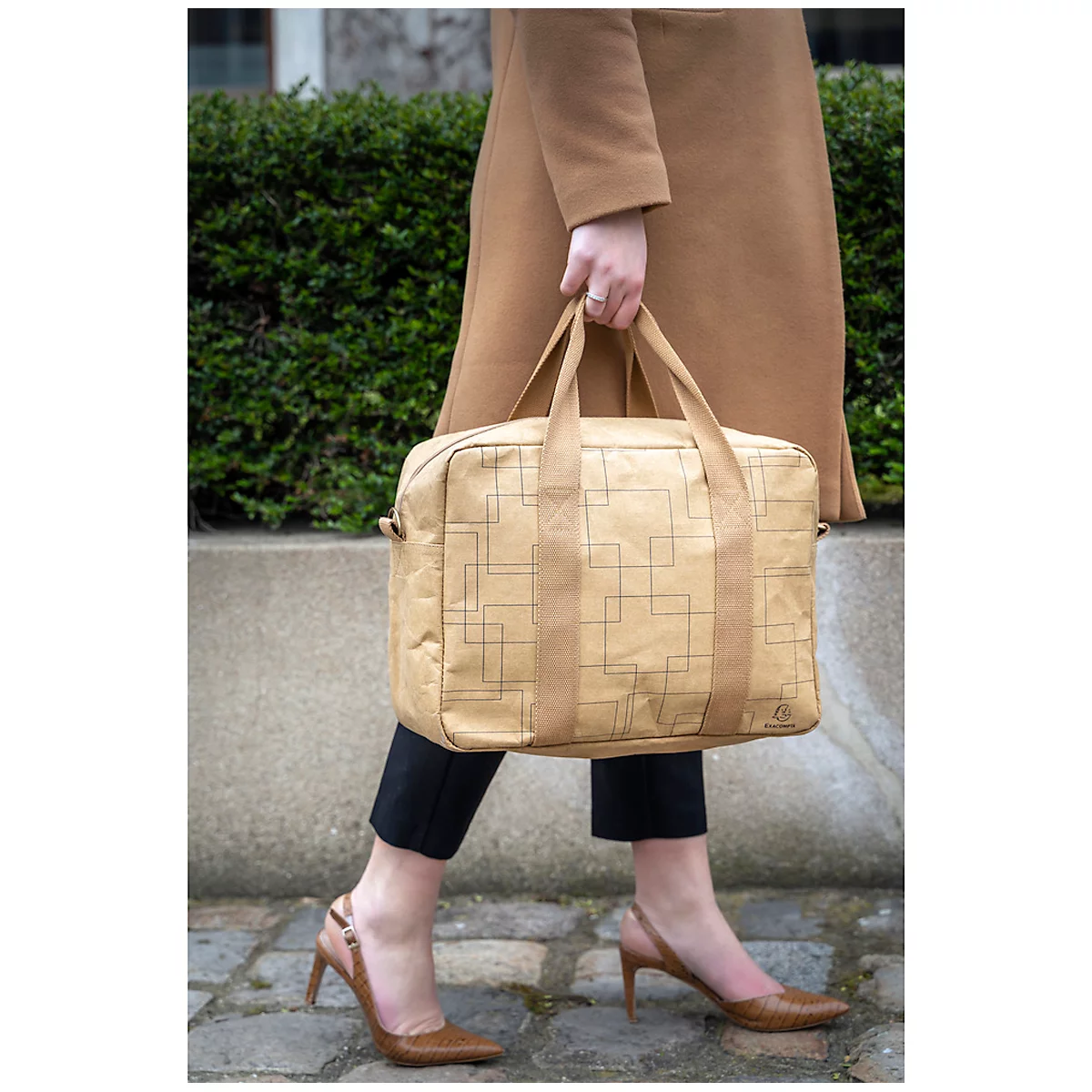 Une personne en manteau marron porte un sac à main beige à motif carré. Elle porte un pantalon noir et des escarpins bruns.