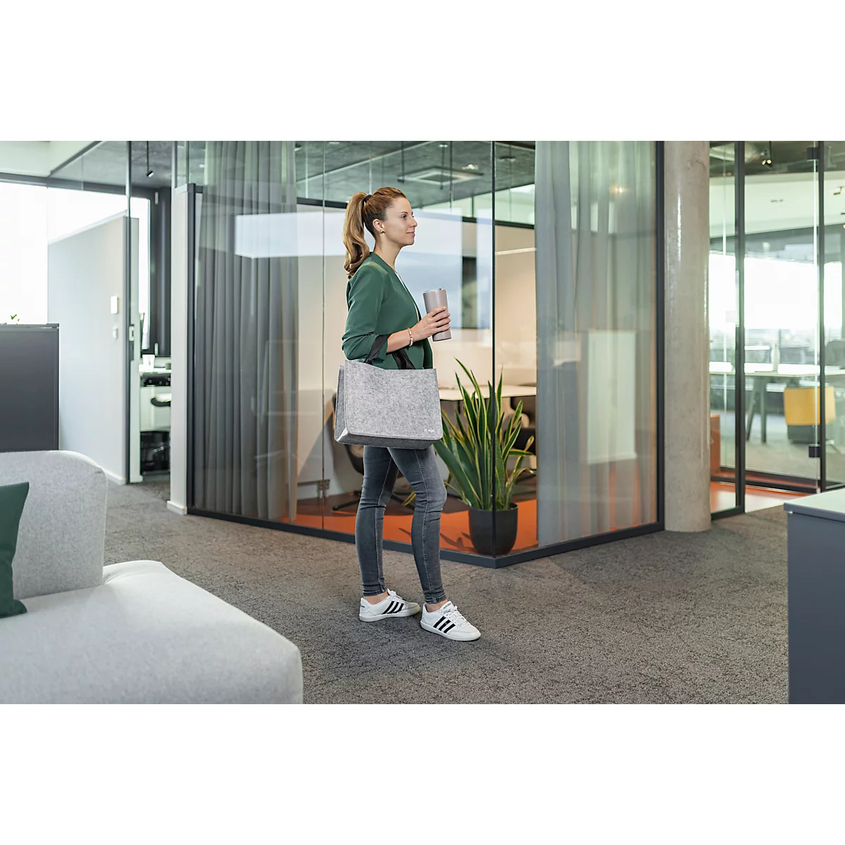 Femme en jean et chemise verte avec sac et tasse marchant dans un bureau. Moquette grise, portes vitrées.