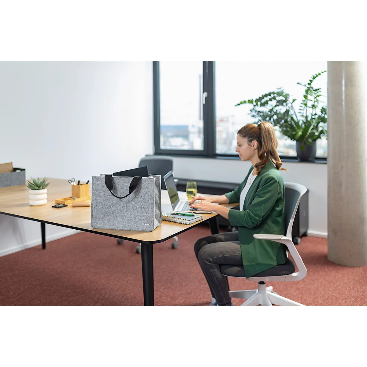 Une femme travaille sur un ordinateur portable au bureau, avec un sac gris à côté d'elle. Elle porte un blazer vert.