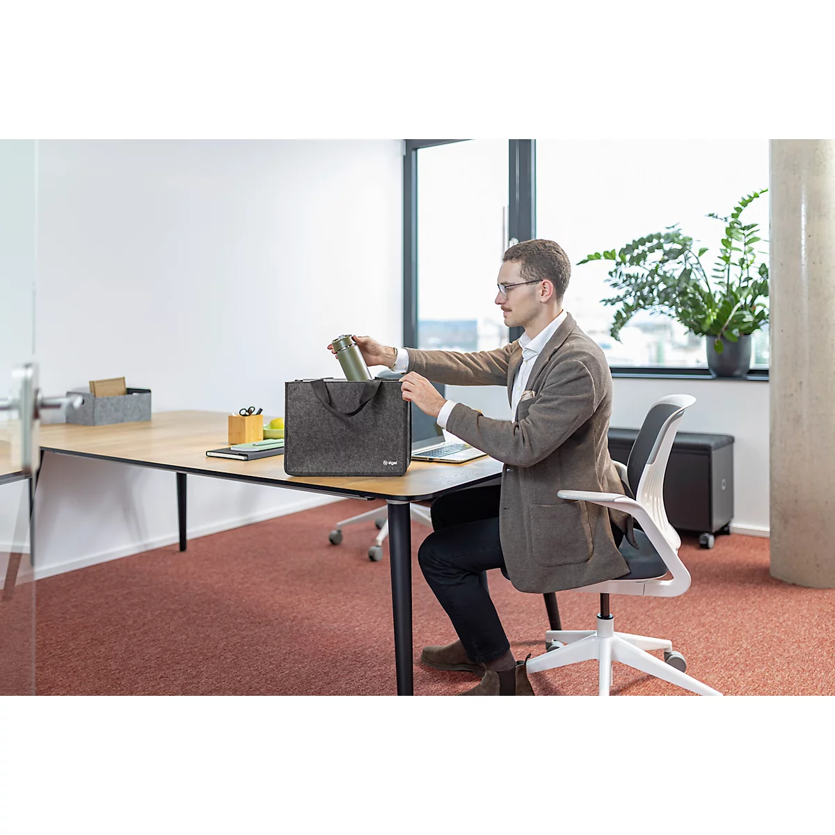 Un homme dans un bureau s'assoit et sort quelque chose d'un sac gris sur le bureau.