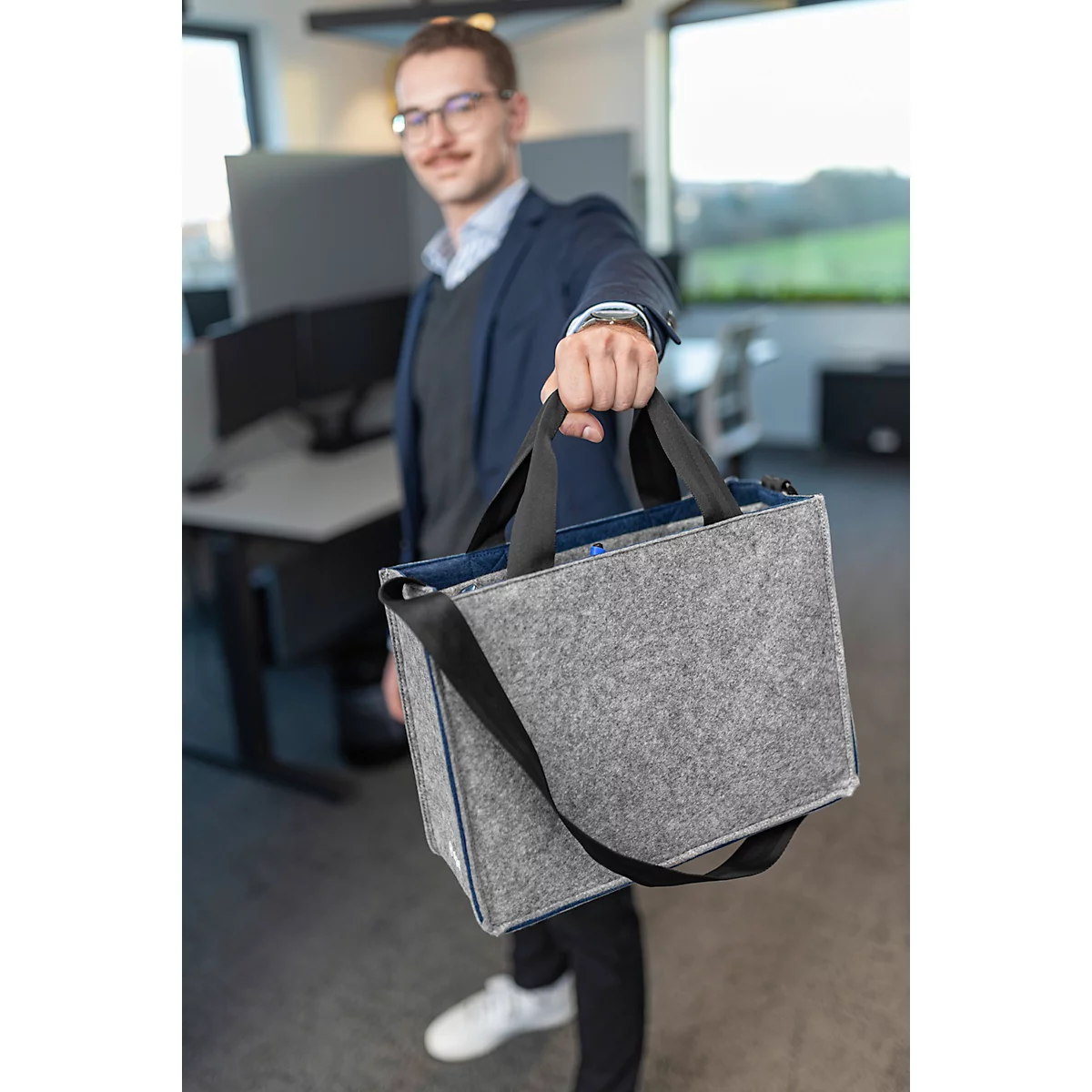 Un homme avec des lunettes présente un sac gris avec une bandoulière noire dans un bureau.