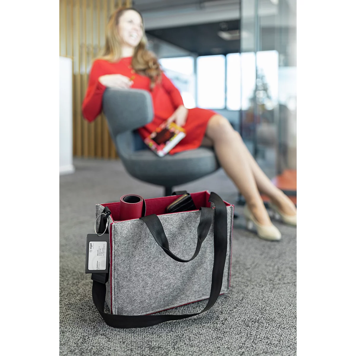 Sac en feutre gris avec doublure rouge, devant une femme souriante en robe rouge. Le sac a une sangle et une poignée noires.