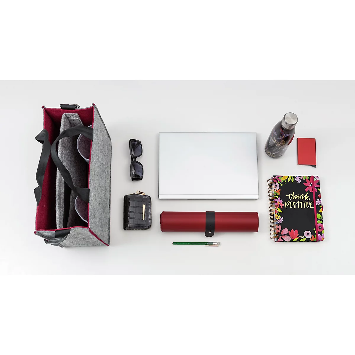 Sur fond clair, un sac gris, des lunettes de soleil, un portefeuille, un ordinateur portable, une bouteille d'eau, un carnet avec "Think Positive" et d'autres accessoires sont disposés.