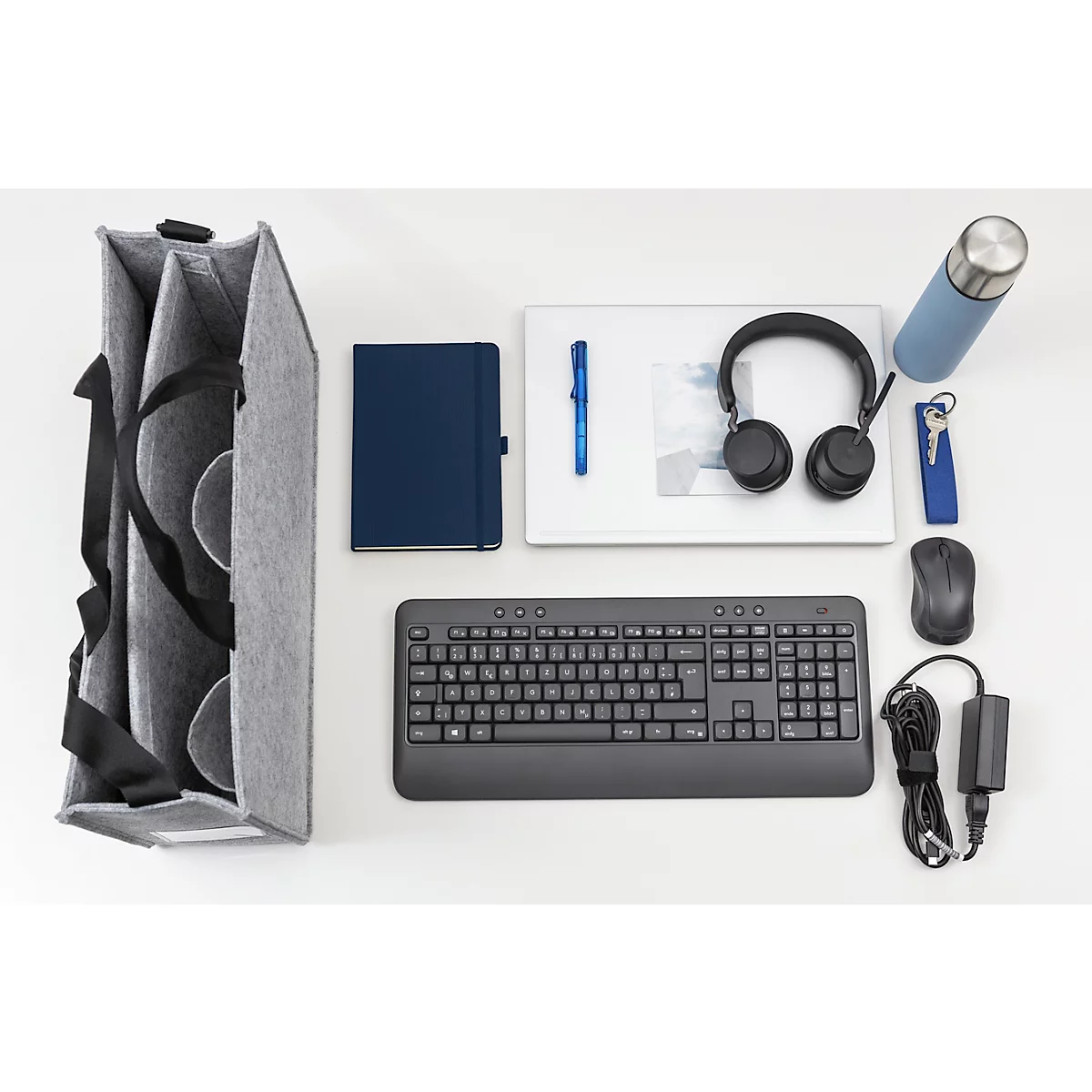 Sac en feutre gris rempli d'une bouteille d'eau, d'un carnet, d'un casque et d'un clavier, posé sur un fond blanc.