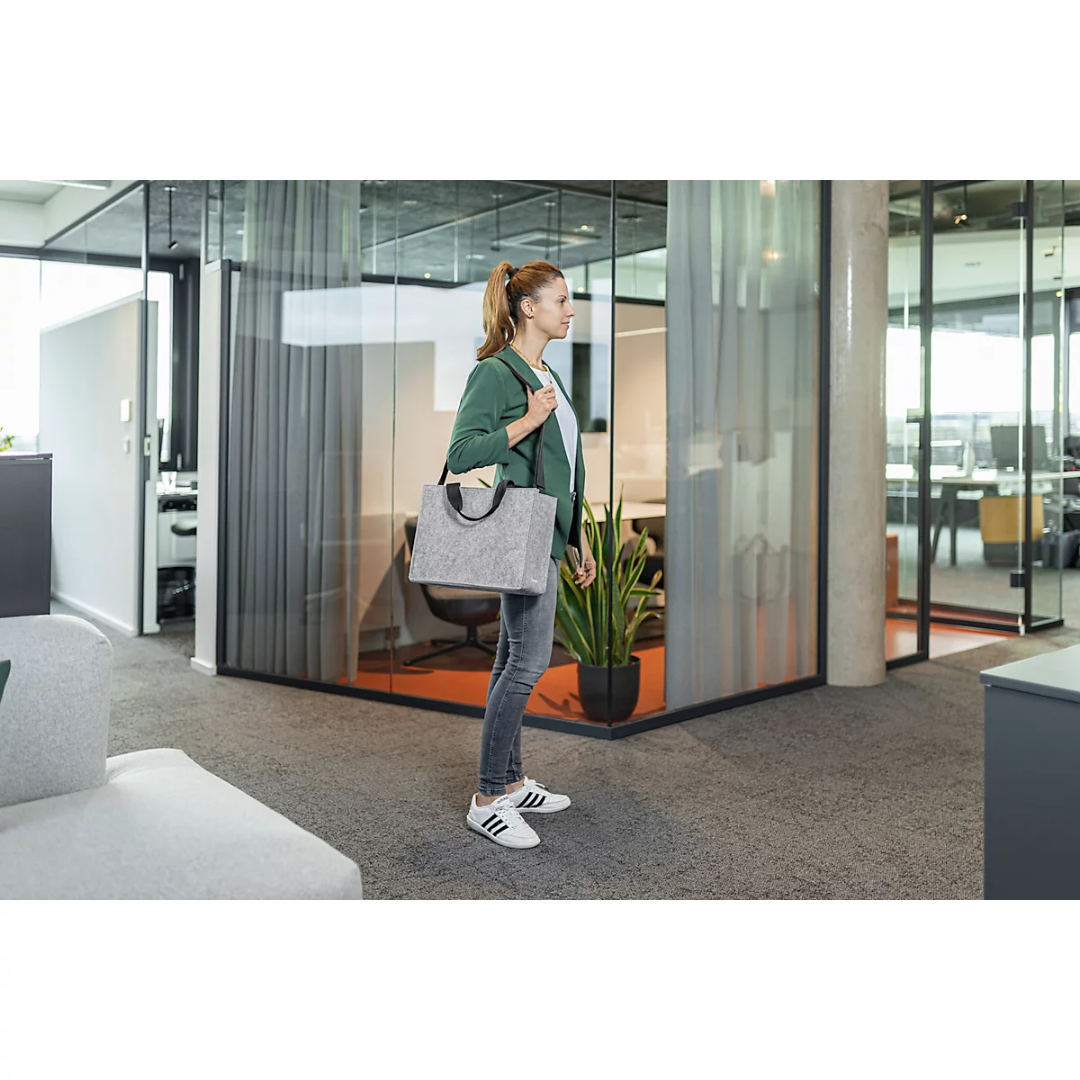 Une femme en jean et blazer vert avec un sac dans un bureau. Elle se tient à côté d'un bureau en verre.