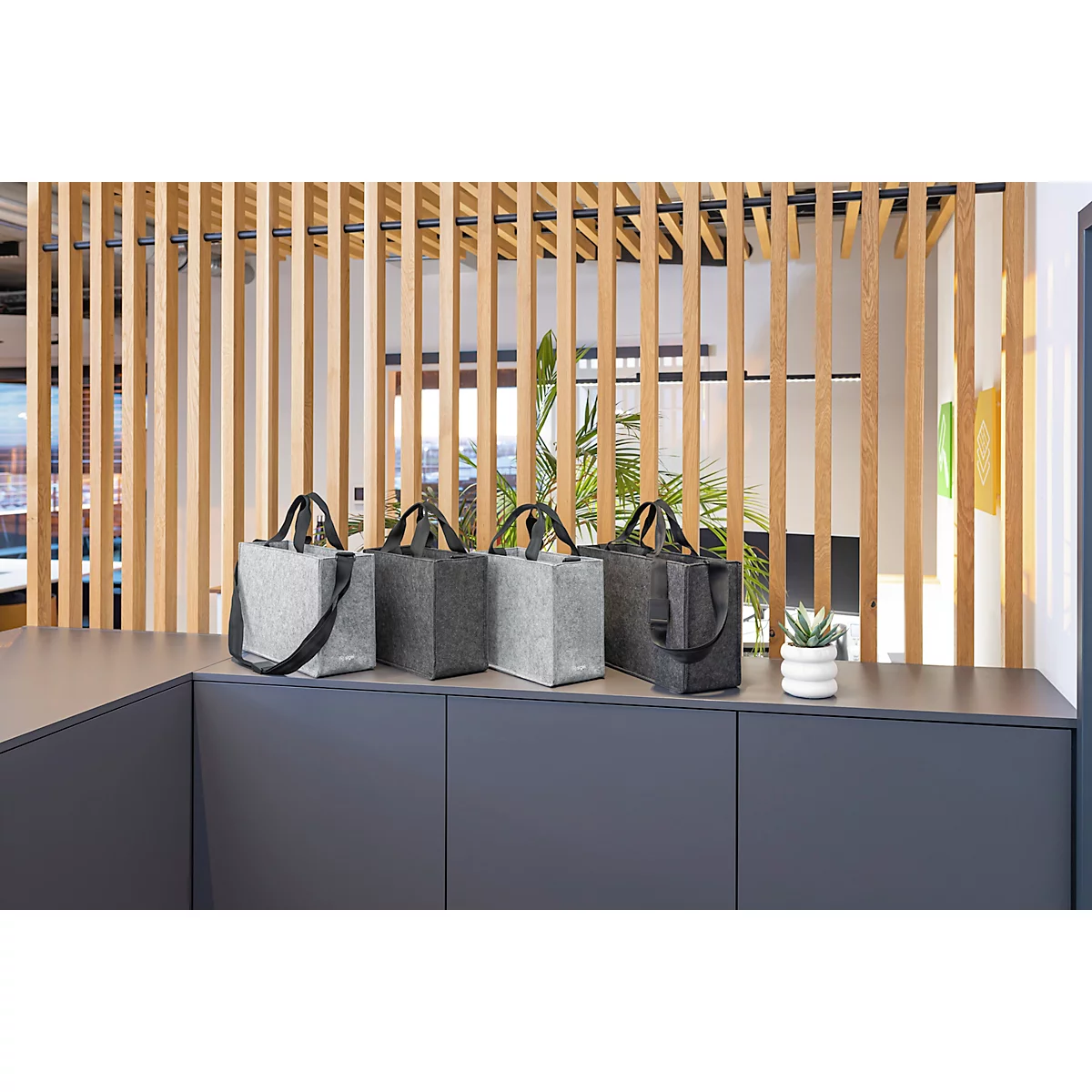 Sacs en feutre gris posés sur une étagère grise devant un mur à lattes en bois. Il y a une petite plante en pot sur l'étagère.
