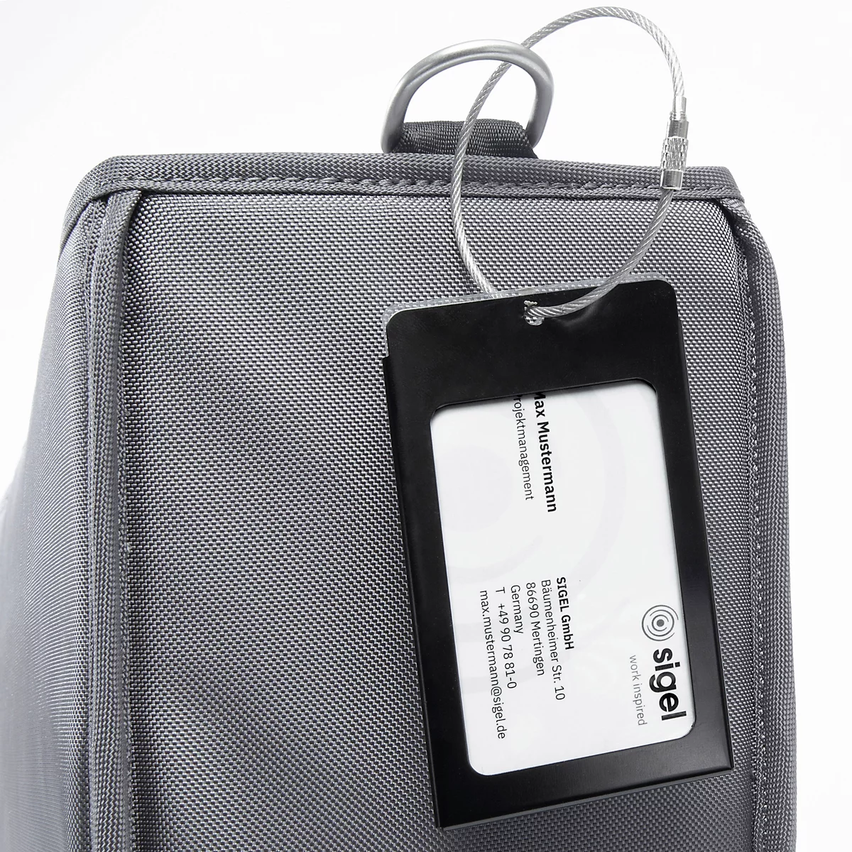 Valise grise avec étiquette nominative. L'étiquette indique le nom, l'adresse et le logo. Les détails sont reconnaissables.