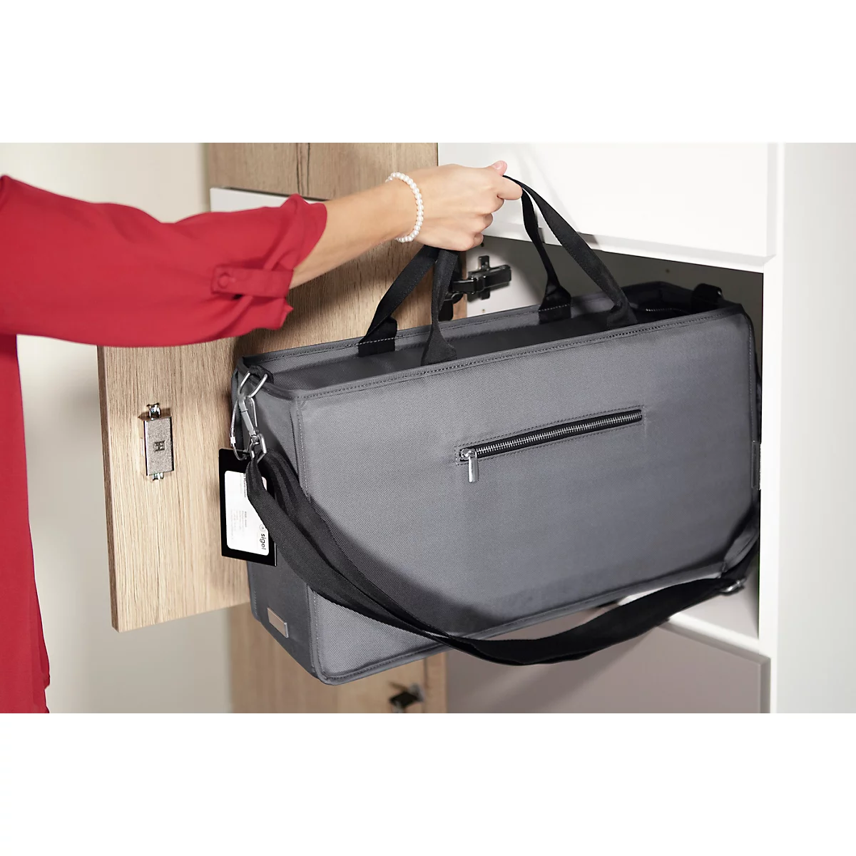Une personne en haut rouge soulève un sac gris hors d'une armoire. Le sac a une bandoulière noire et une fermeture éclair.