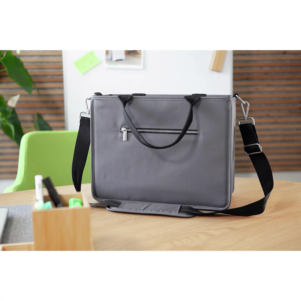 Sacoche d'ordinateur portable grise avec bandoulière noire sur une table en bois. En arrière-plan, une chaise verte et des fournitures de bureau.