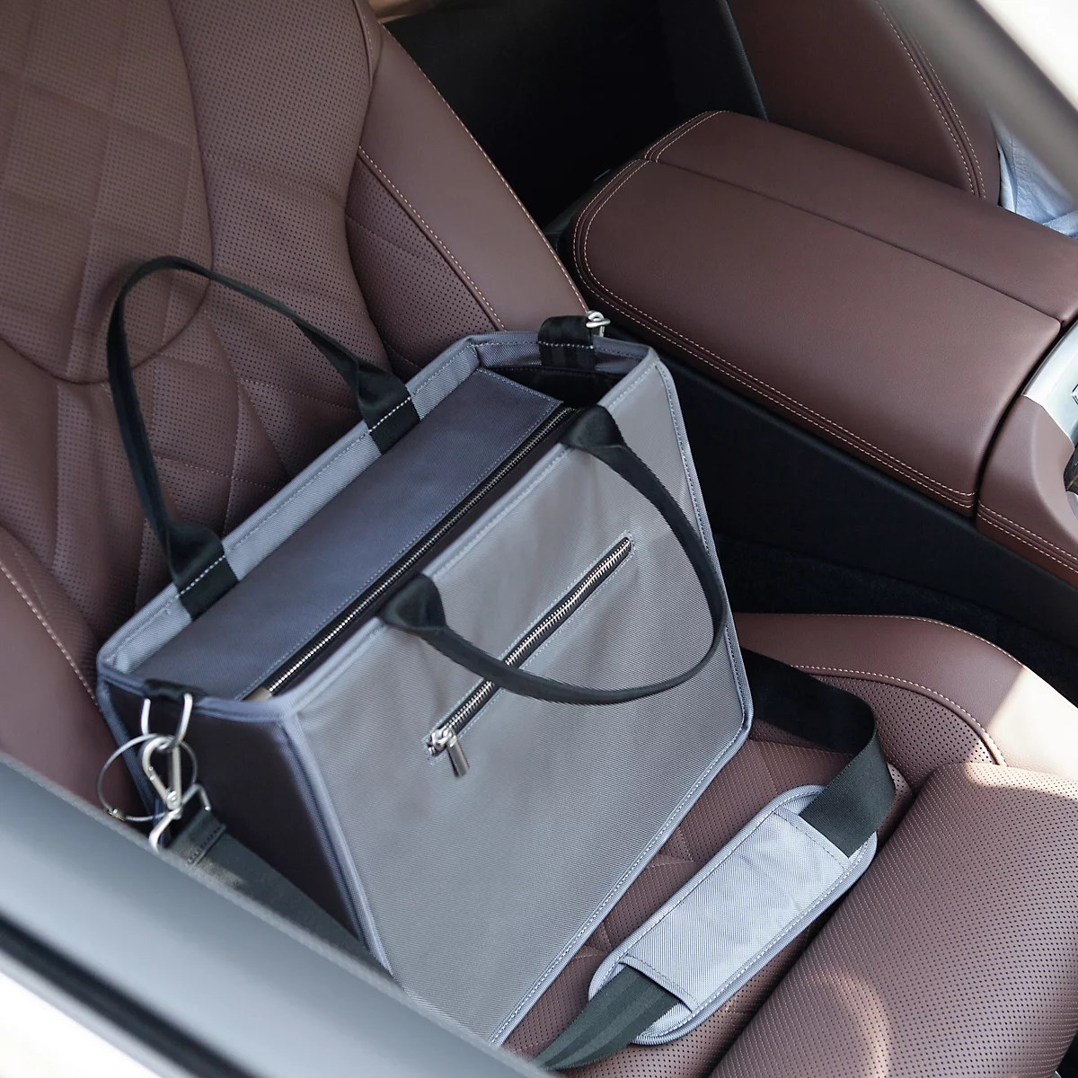 Sac à main gris avec une sangle de transport noire sur un siège de voiture marron. Le sac a une fermeture éclair et une bandoulière supplémentaire.